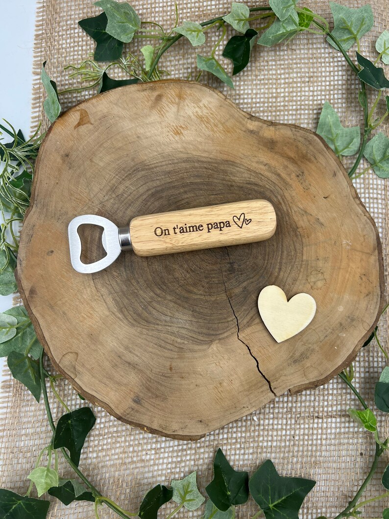 May include: A bottle opener with a light-colored wooden handle featuring the engraved text On t'aime papa and a small heart symbol, accompanied by a separate small wooden heart shape, both resting on a natural wood slice surface.