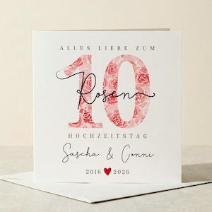 Könnte beinhalten: Eine weiße Grußkarte mit dem Text "ALLES LIEBE ZUM 10 ROSEN HOCHZEITSTAG Sascha & Conni 2016 ❤️ 2026". Die Zahl 10 ist mit einem rosa Blumenmuster gefüllt. Die Karte liegt auf einem weißen Umschlag.