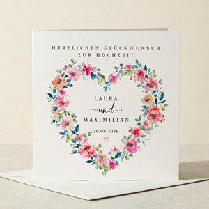 Könnte beinhalten: Eine weiße Hochzeitskarte mit einem floralen Herzdesign. Das Herz besteht aus rosa, orangefarbenen und grünen Aquarellblumen. Die Karte enthält die Namen "Laura" und "Maximilian" sowie das Datum "26.09.2026".
