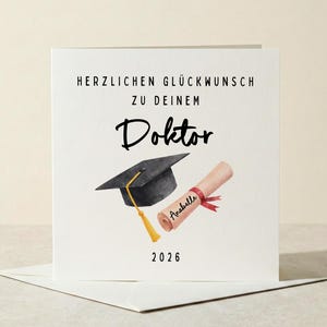 Peut inclure: Carte de félicitations de graduation blanche avec le texte allemand "HERZLICHEN GLÜCKWUNSCH ZU DEINEM DOKTOR" et l'année 2026. La carte présente une toque noire avec un gland doré et un diplôme roulé avec un ruban rouge.