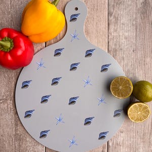 May include: A gray, round cutting board with a handle, decorated with blue seashells and starfish. Yellow and red bell peppers, along with halved lemons, are arranged around the board on a wooden surface.
