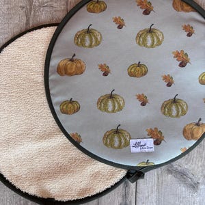 May include: Two round pot holders with a light grey top surface featuring a repeating pattern of orange and green pumpkins and brown oak leaves. The underside is a textured beige fabric. A small white label reads Lilliput Home Designs.