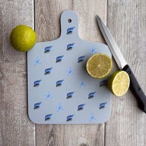 May include: A light blue cutting board with a handle, decorated with seashell and starfish patterns. A lime and a halved lime are next to the board, along with a knife. The board is on a wooden surface.