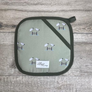 以下が含まれることがあります： 緑色の鍋つかみで、白い羊の模様と緑色の布製のループが付いています。鍋つかみには「Lilliput Home Designs」という文字が印刷されています。