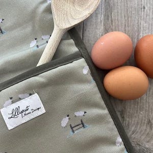 Puede incluir: Un delantal verde salvia con un estampado de ovejas y una cuchara de madera. El delantal tiene un ribete verde oscuro y una etiqueta que dice "Lilliput Home Designs". Tres huevos marrones están junto al delantal.