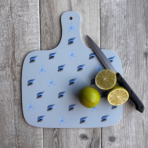 May include: A light blue cutting board with a handle, decorated with blue seashells and starfish. A whole lime and a lime cut in half are next to a black-handled knife. The board rests on a wooden surface.