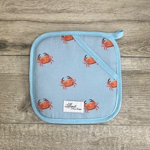 Puede incluir: Manopla azul con un patrón de cangrejos naranjas. La manopla tiene un bolsillo en la parte posterior y dice "Lilliput Home Designs" en la esquina inferior derecha.