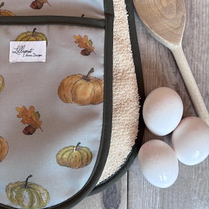 Puede incluir: Un paño de cocina gris claro con estampado de calabazas y hojas de otoño, con ribete verde oscuro y reverso de felpa blanca. Una etiqueta blanca dice Lilliput Home Designs. Tres huevos blancos y una cuchara de madera junto al paño.