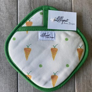Puede incluir: Un agarrador de olla verde y blanco con un patrón de zanahorias y lunares verdes. El agarrador de olla tiene un borde verde y el texto "Lilliput Home Designs" está impreso en la etiqueta.
