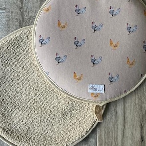 May include: Two round kitchen trivets. One is beige with a textured surface. The other is a light tan colour with a pattern of chickens in blue, grey, and yellow. A small white tag with text is visible.