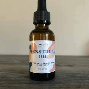 May include: A brown glass bottle with a black dropper top. The label is white with pink and blue abstract shapes and the text "PNEUMA BODY & WELLNESS MENSTRUAL OIL WE'LL GET THROUGH THIS TOGETHER! 1 fl oz (30 ml)"