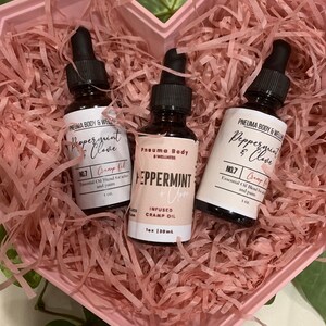May include: Two small brown glass bottles with dropper tops, filled with essential oil blends. The bottles are labelled "Peppermint & Clove" and "Peppermint + Clove Infused Cramp Oil". The bottles are in a pink heart-shaped box with pink crinkle paper.