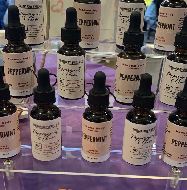 May include: A display of small glass bottles filled with essential oil blends. The bottles are labelled with the text "Peppermint & Clove" and "Peppermint" and have a black dropper cap. The bottles are arranged on a clear acrylic shelf.