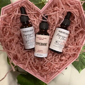 May include: Three small glass bottles with dropper tops filled with essential oil. The bottles are labelled "Peppermint & Clove" and "Peppermint" with the brand name "Pneuma Body & Wellness". The bottles are in a pink heart-shaped box filled with pink crinkle paper.