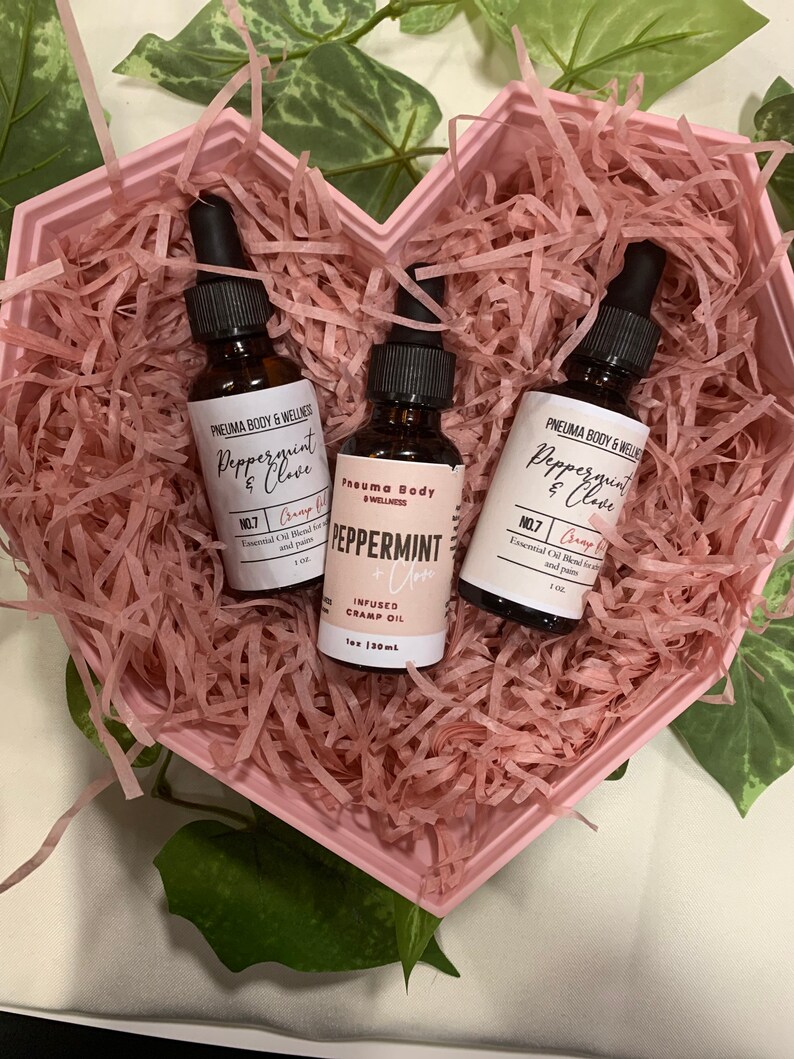 May include: Three small glass bottles with dropper tops filled with essential oil. The bottles are labelled "Peppermint & Clove" and "Peppermint" with the brand name "Pneuma Body & Wellness". The bottles are in a pink heart-shaped box filled with pink crinkle paper.