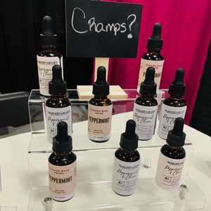 May include: A display of eight small glass bottles with dropper tops, filled with a brown liquid. The bottles are labelled "Peppermint" and "Peppermint & Clove" with the brand name "Pneuma Body & Wellness". The bottles are arranged on a clear acrylic shelf with a black chalkboard sign that says "Champs?" above the bottles.