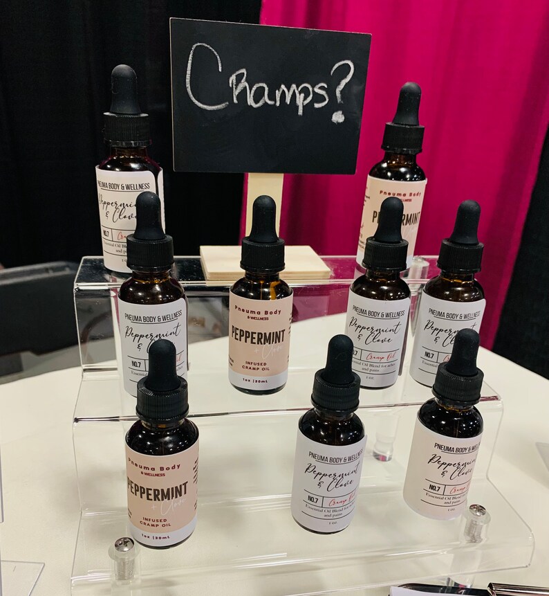 May include: A display of eight small glass bottles with dropper tops, filled with a brown liquid. The bottles are labelled "Peppermint" and "Peppermint & Clove" with the brand name "Pneuma Body & Wellness". The bottles are arranged on a clear acrylic shelf with a black chalkboard sign that says "Champs?" above the bottles.