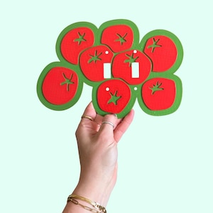 May include: A decorative light switch cover featuring a cluster of red tomatoes with green leaves. The tomatoes have white highlights and are arranged on a green background. The cover is held up by a hand.