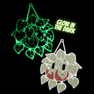 May include: Two decorative light switch covers in the shape of hanging plants. One glows green in the dark, and the other is white with green leaves and red accents. The text "GLOW IN THE DARK" is also visible.