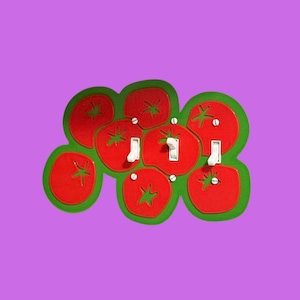May include: A triple light switch wall plate featuring a cluster of red tomato shapes with green outlines. The plate includes three white toggle switches positioned within the tomato design, secured by visible screws on a solid purple background.