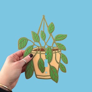 May include: A decorative light switch cover featuring a potted plant design. The cover is painted with a light tan pot, green leaves, and a gold-colored hanging structure. The background is a solid light blue.