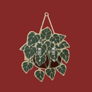 May include: Decorative light switch cover featuring a hanging plant design. The cover has a cream-colored frame with green leaves and a brown pot. Two silver light switches are visible. The background is a solid red.