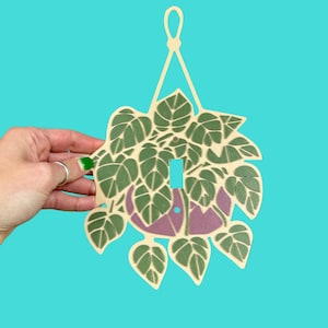 May include: A decorative light switch cover featuring a plant design. The cover is cream-colored with green leaves and a purple pot. The switch opening is rectangular. The cover is held against a turquoise background.