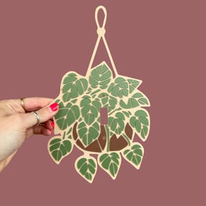 May include: A decorative light switch cover featuring a hanging plant design. The cover is made of light beige material with green leaves and a brown pot. The switch opening is a rectangular shape. The background is a solid mauve color.