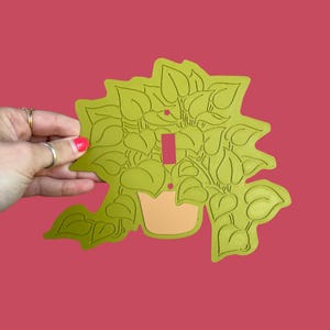 May include: A decorative light switch cover featuring a green plant design. The cover has a light green background with a detailed outline of leaves and a pot. The switch opening is a rectangular shape in a pink color.