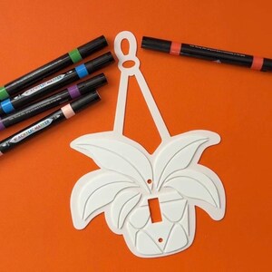 May include: White decorative light switch cover in the shape of a potted plant, with several black acrylic markers with colored ends. The background is a solid orange color.