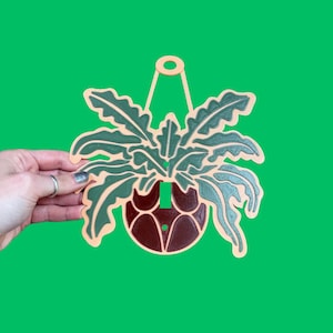 May include: A wooden wall hanging featuring a stylized plant in a pot. The plant has green leaves with light green and beige accents. The pot is brown, and the background is green. The wall hanging is designed to be hung from a loop at the top.