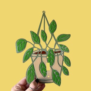 May include: A decorative light switch cover in the shape of a hanging plant. The cover features a beige pot with green leaves and a green hanging structure. The background is a solid yellow.