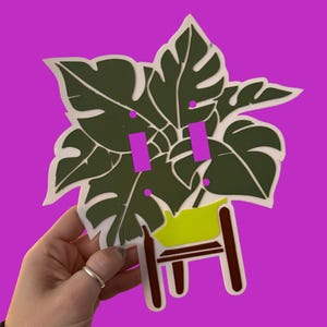 May include: A decorative light switch cover featuring a green monstera leaf design with white outlines. The cover has two pink switch toggles and a small green and brown plant stand. The background is a solid purple.
