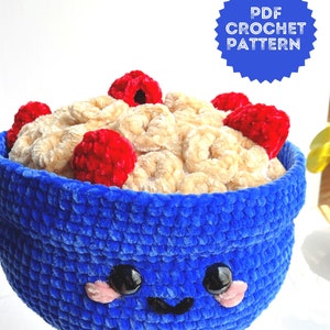 Pode incluir: Uma tigela azul de crochê com um rosto feliz e olhos pretos. A tigela está cheia de cereais bege de crochê e framboesas vermelhas.