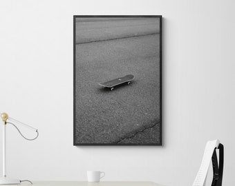 May include: Black and white photograph of a skateboard on a paved road. The photo is framed in a black frame and is hanging on a white wall.