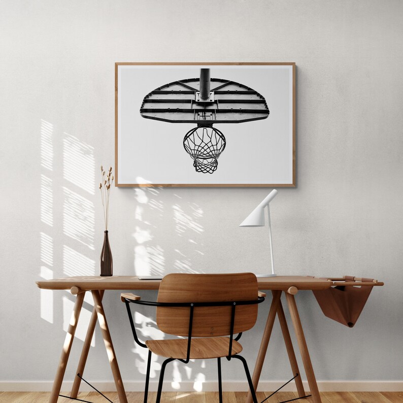 May include: A black and white photograph of a basketball hoop hanging from the ceiling. The hoop is in focus, while the background is blurred. The photo is framed in a wooden frame.