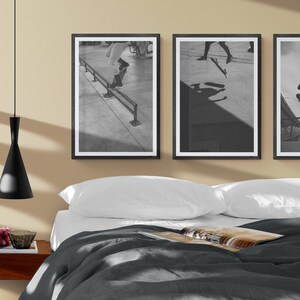 Puede incluir: Tres fotograf&iacute;as enmarcadas en negro de patinadores haciendo trucos en una rampa. Las fotograf&iacute;as est&aacute;n colgadas en una pared beige sobre una cama con ropa de cama blanca y un edred&oacute;n gris.