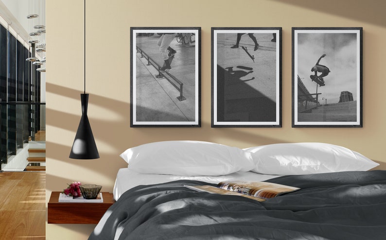 Puede incluir: Tres fotograf&iacute;as enmarcadas en negro de patinadores haciendo trucos en una rampa. Las fotograf&iacute;as est&aacute;n colgadas en una pared beige sobre una cama con ropa de cama blanca y un edred&oacute;n gris.
