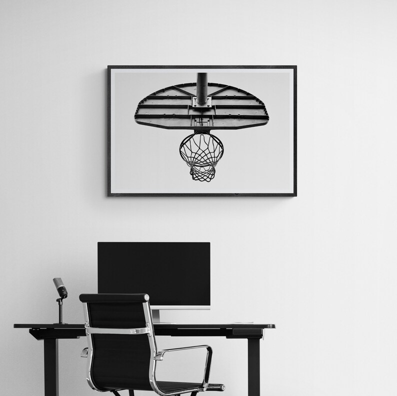 May include: Black and white photograph of a basketball hoop hanging from a ceiling. The hoop is made of metal and has a net. The photo is framed and hanging on a white wall.