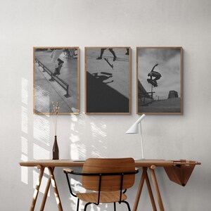 Puede incluir: Tres fotograf&iacute;as en blanco y negro enmarcadas de patinadores haciendo trucos. Las fotos est&aacute;n colgadas en una pared blanca sobre un escritorio de madera con una silla.