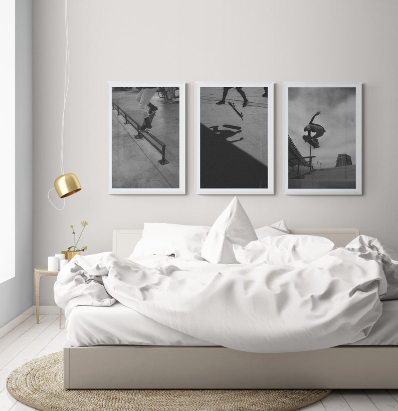 Puede incluir: Tres fotograf&iacute;as en blanco y negro de patinadores haciendo trucos. Las fotos est&aacute;n enmarcadas en blanco y colgadas en una pared blanca sobre una cama blanca con ropa de cama blanca.
