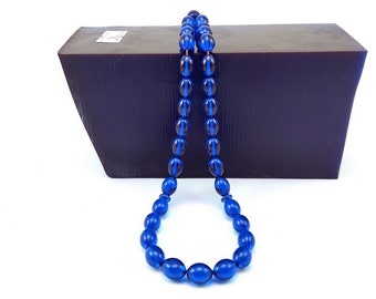 May include: A blue beaded necklace with oval and round beads. The necklace is displayed in front of a dark brown wooden block. The beads are a translucent blue color, and the necklace is hanging in a loop.