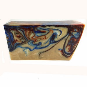 May include: A rectangular soap bar with a marbled pattern. The soap has swirls of blue, red, and cream colours. The top edge has a dark brown hue, and the soap is set against a white background.