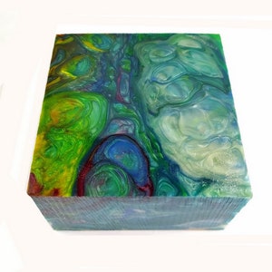 May include: A square block with a colorful, swirled design. The top surface features a mix of green, blue, yellow, and red hues, resembling a marbled effect. The sides of the block have a textured appearance, with a predominantly blue color.