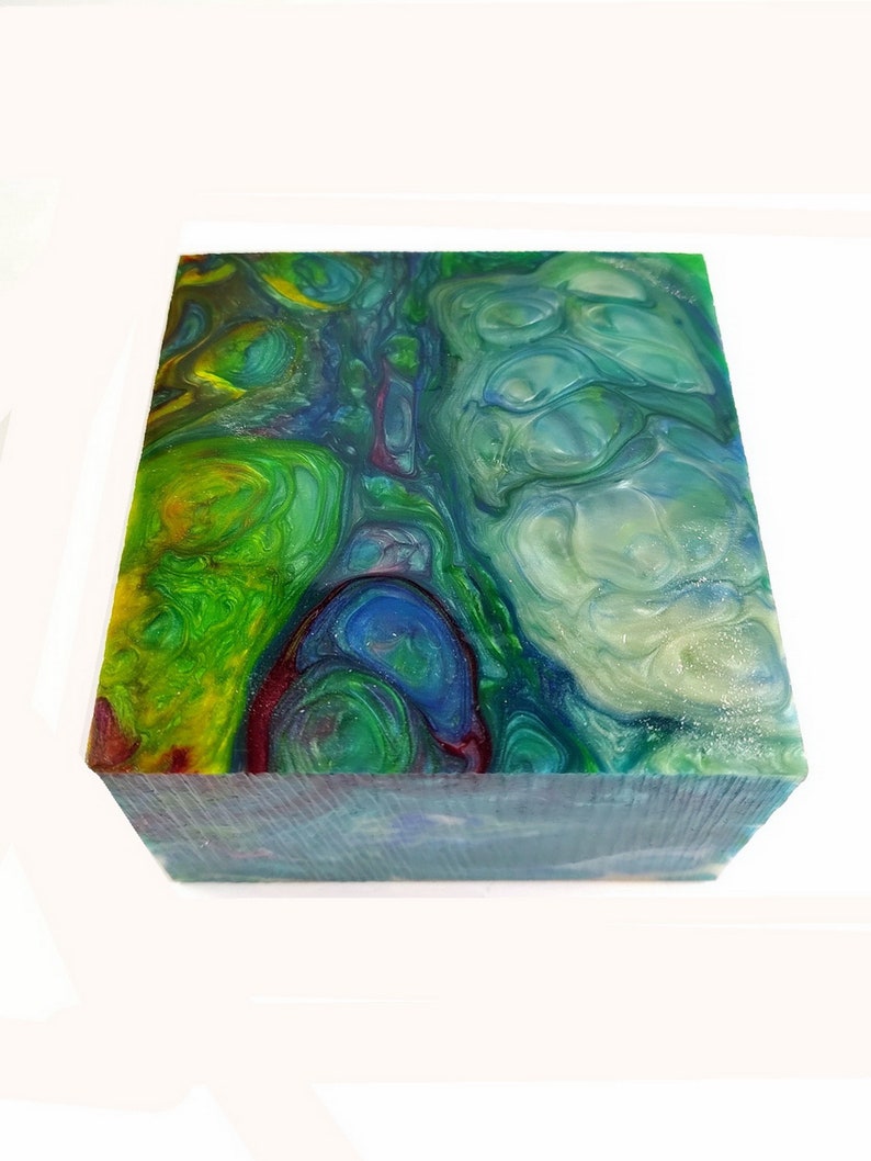 May include: A square block with a colorful, swirled design. The top surface features a mix of green, blue, yellow, and red hues, resembling a marbled effect. The sides of the block have a textured appearance, with a predominantly blue color.