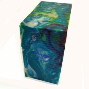 May include: A rectangular block of swirled resin in shades of teal, blue, green, and cream. The block has a polished surface with visible layers and a marbled effect. The colors blend together, creating a unique and artistic appearance.