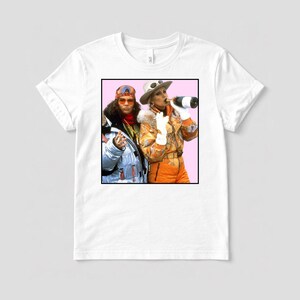 Könnte beinhalten: Weißes T-Shirt mit einem gerahmten Bild von zwei Personen in Skianzügen. Eine Person trinkt Champagner, die andere trägt ein Stirnband und eine Sonnenbrille. Das Bild hat einen rosa Hintergrund.