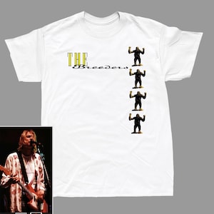 Könnte beinhalten: Weißes T-Shirt mit dem Bandnamen "The Breeders" in gelbem und schwarzem Text. Vier kleine Affenfiguren sind vertikal auf der rechten Seite ausgerichtet. Ein kleines Bild einer Person, die Gitarre spielt, befindet sich unten links.
