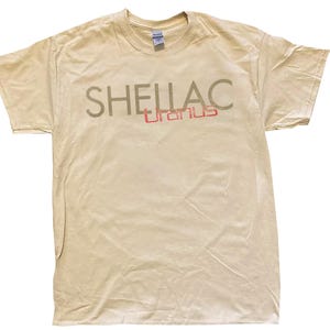 Könnte beinhalten: Ein hellbeiges Kurzarm-T-Shirt mit dem Wort SHELLAC in großen, dunkelolivfarbenen Großbuchstaben auf der Brust und dem darunter gedruckten Wort Uranus in einer kleineren, rot umrandeten Schriftart. Das Shirt hat einen klassischen Rundhalsausschnitt.