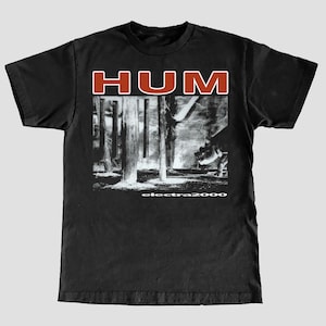 May include: Black t-shirt with the band name "HUM" in red block letters at the top. Below is a black and white image of a scene with wooden posts. The text "electra2000" is at the bottom of the t-shirt.